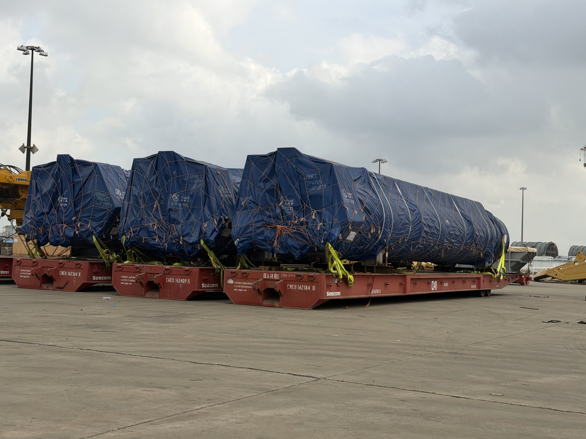 Three long loads covered in blue tarpaulins and rope netting, lashed to red flat racks in an open paved yard under an overcast sky.